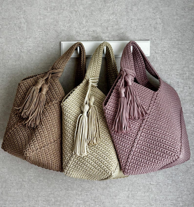 May include: Three crochet tote bags in shades of brown, beige, and mauve hang against a gray wall. Each bag features a knotted handle and decorative tassels. The bags are made of a textured, woven material.