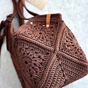 May include: A dark brown crochet bag constructed from multiple square motifs featuring a central sunburst pattern. The bag includes a matching crocheted shoulder strap and a small tan leather tag with the text LUNAR CA attached to the upper edge.
