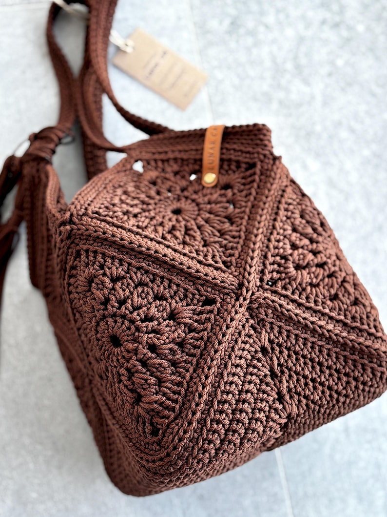 May include: A dark brown crochet bag constructed from multiple square motifs featuring a central sunburst pattern. The bag includes a matching crocheted shoulder strap and a small tan leather tag with the text LUNAR CA attached to the upper edge.