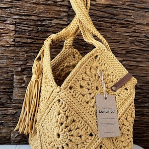 May include: A handmade, mustard-yellow crochet bag with a geometric pattern. The bag features a long handle, a tassel, and a tag that reads "Lunar cat". The bag is made of a soft, textured yarn.