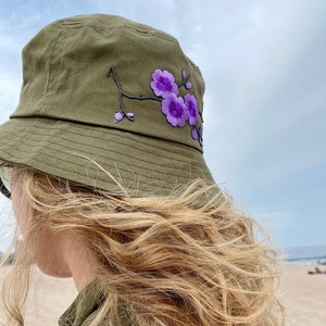 May include: A green bucket hat with a purple embroidered flower design.