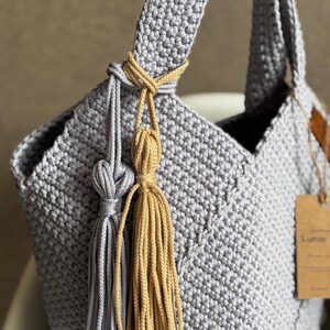 May include: A handmade gray crochet tote bag with a knotted handle and decorative tassels in gray and gold. The bag features a textured stitch pattern and a small tag. The bag is designed for carrying items.