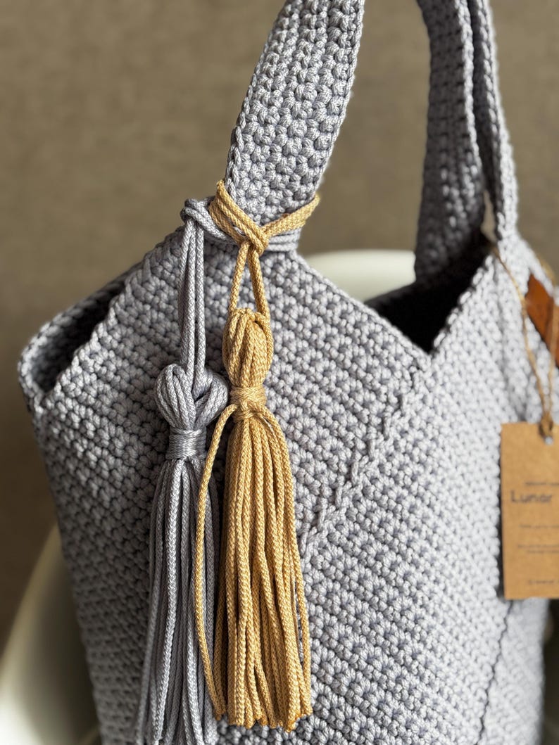 May include: A handmade gray crochet tote bag with a knotted handle and decorative tassels in gray and gold. The bag features a textured stitch pattern and a small tag. The bag is designed for carrying items.