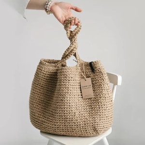 Puede incluir: Un bolso tote de yute tejido con un asa trenzada. El bolso es de color beige natural y tiene una etiqueta que dice "Lunar cat". El bolso est&aacute; sobre una silla blanca.