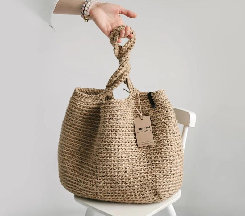 Puede incluir: Un bolso tote de yute tejido con un asa trenzada. El bolso es de color beige natural y tiene una etiqueta que dice "Lunar cat". El bolso est&aacute; sobre una silla blanca.