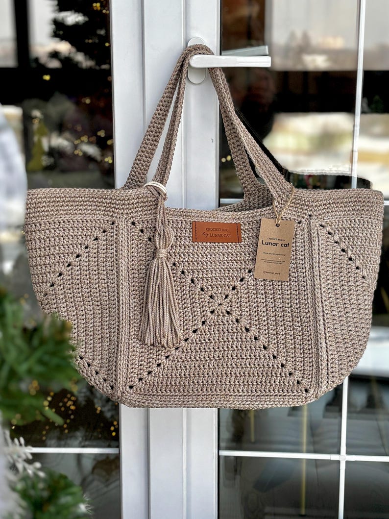 May include: A beige crochet tote bag with a geometric design. The bag has two handles and a decorative tassel. A small leather tag reads "Crochet Bag by Lunar Cat" and a paper tag reads "Lunar Cat".