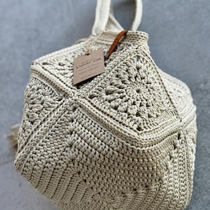 May include: A beige crochet handbag with a geometric, multi-faceted structure. The bag features intricate star-like patterns on its panels and a single top handle. A brown rectangular paper tag with the text Lunar cat is attached by a string.