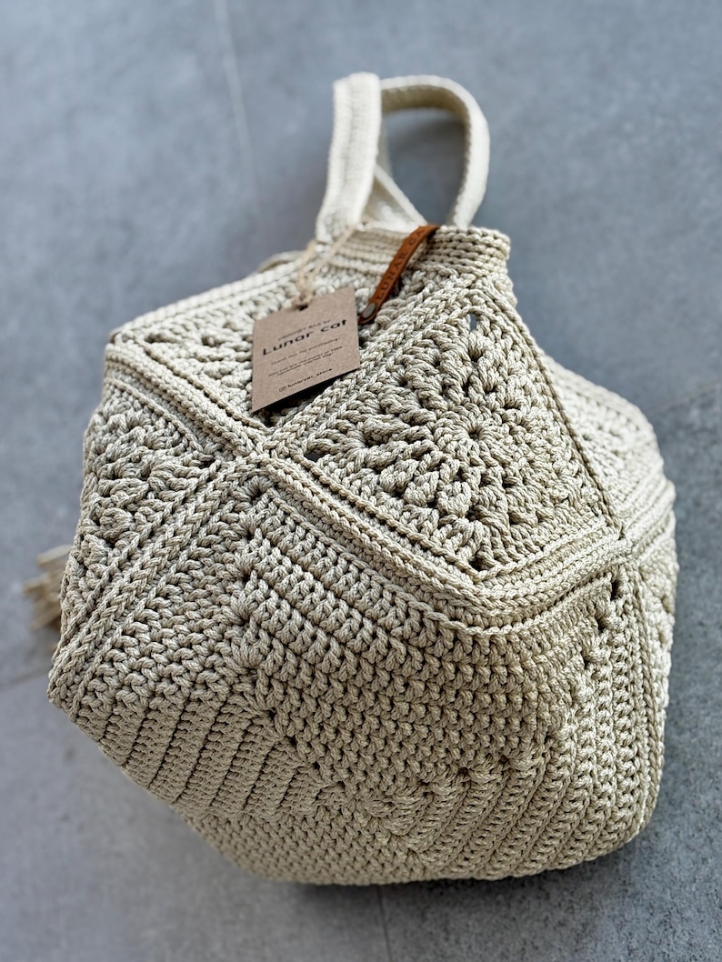 May include: A beige crochet handbag with a geometric, multi-faceted structure. The bag features intricate star-like patterns on its panels and a single top handle. A brown rectangular paper tag with the text Lunar cat is attached by a string.