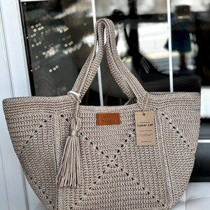 May include: A beige crocheted tote bag with a geometric pattern and a tassel. The bag has two handles and a leather tag that says "by Lunar cat". A paper tag with the words "Lunar cat" is attached to the bag.