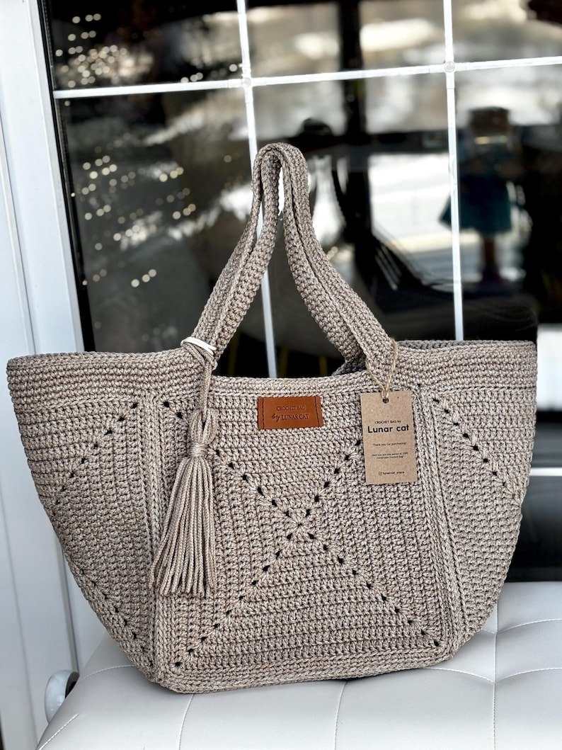May include: A beige crocheted tote bag with a geometric pattern and a tassel. The bag has two handles and a leather tag that says "by Lunar cat". A paper tag with the words "Lunar cat" is attached to the bag.