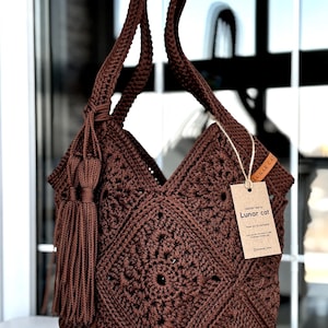 May include: A dark brown crochet bag featuring a square motif pattern, a single shoulder strap, and a decorative tassel. A rectangular cardboard tag attached with twine displays the text Lunar cat and additional printed information.