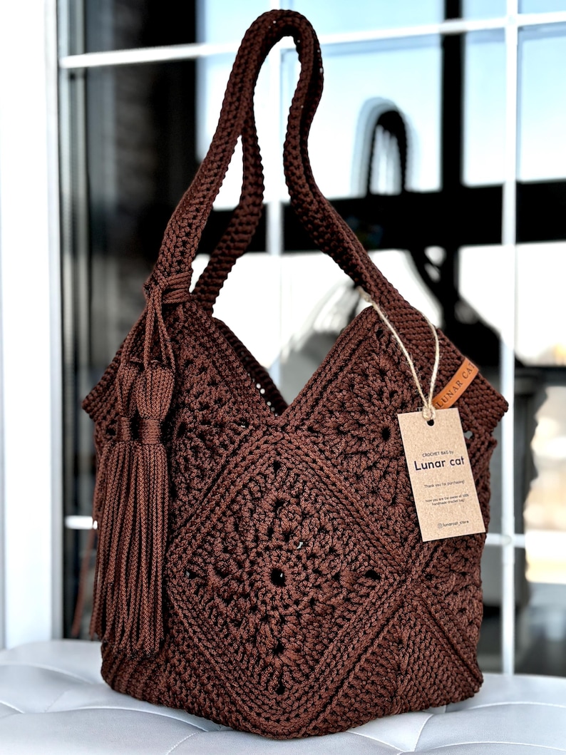 May include: A dark brown crochet bag featuring a square motif pattern, a single shoulder strap, and a decorative tassel. A rectangular cardboard tag attached with twine displays the text Lunar cat and additional printed information.