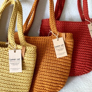 May include: Three crocheted tote bags in yellow, orange, and red. Each bag has a leather tag with the text "Lunar cat" and a website address. The bags are handmade and made of 100% cotton.