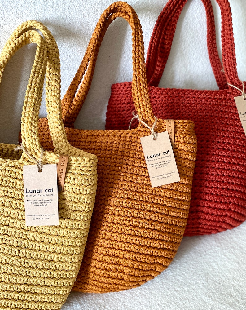 May include: Three crocheted tote bags in yellow, orange, and red. Each bag has a leather tag with the text "Lunar cat" and a website address. The bags are handmade and made of 100% cotton.