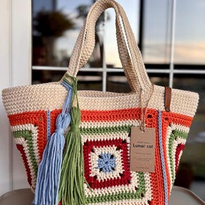 May include: A crochet tote bag featuring a granny square pattern in cream, orange, green, red, and light blue. The bag has two handles, a light blue tassel, a green tassel, and a brown paper tag with the text Lunar cat attached by twine.