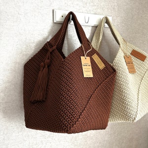 May include: Two crocheted tote bags hanging on a hook. The bag on the left is brown with a brown tassel and a tag that says "Lunar cel." The bag on the right is beige with a tag that says "Lunar cel."