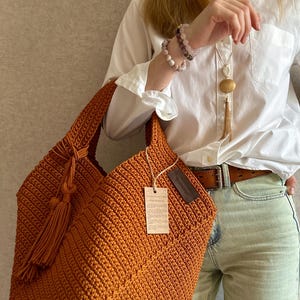 May include: A handcrafted, orange crochet tote bag with a tassel detail. The bag features a textured stitch pattern and a tag attached to the side. The bag is held by a person wearing a white shirt and light-colored jeans.
