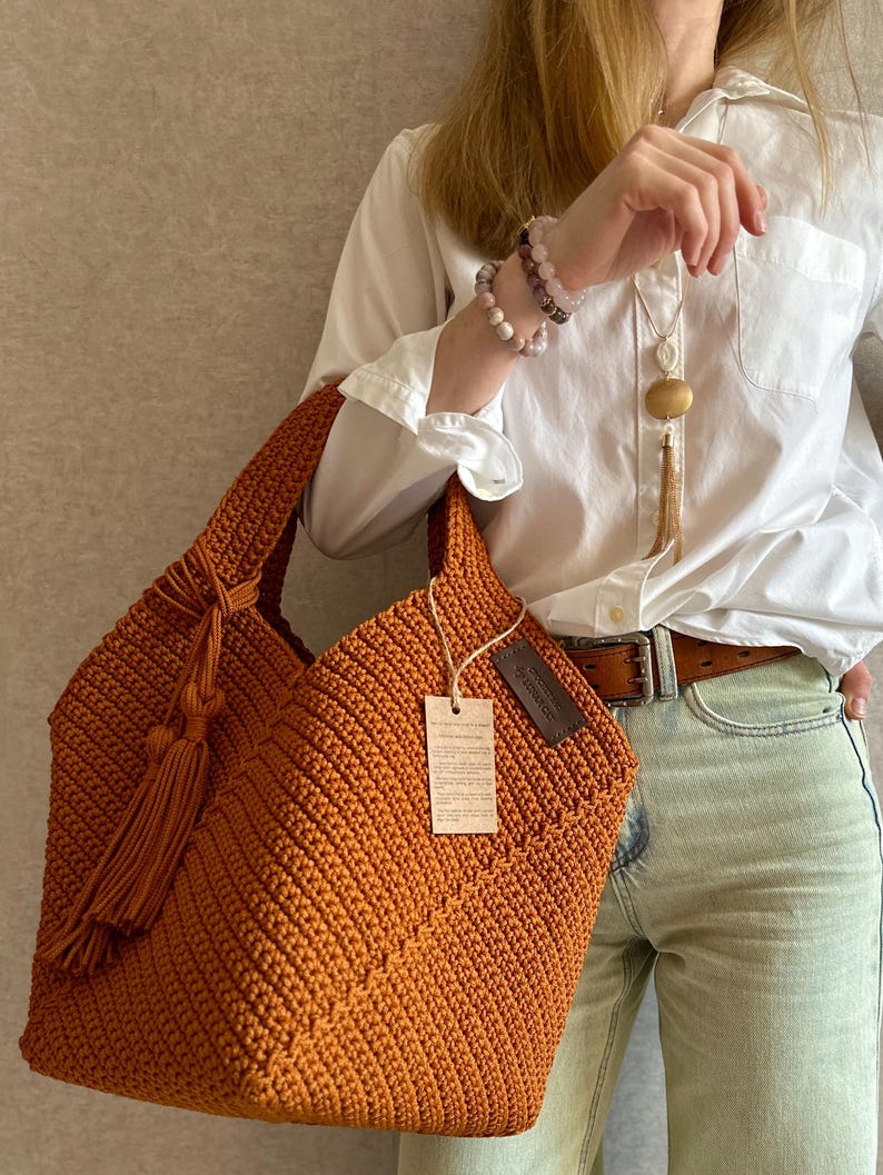 May include: A handcrafted, orange crochet tote bag with a tassel detail. The bag features a textured stitch pattern and a tag attached to the side. The bag is held by a person wearing a white shirt and light-colored jeans.