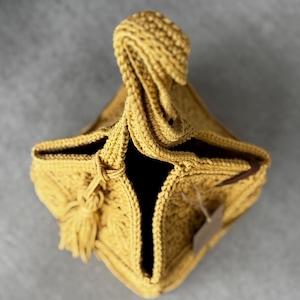 May include: Mustard yellow crocheted bag with a knotted handle and a tassel. The bag has a square base and a dark interior. A small tag is attached to the side. The bag is made of a textured yarn.