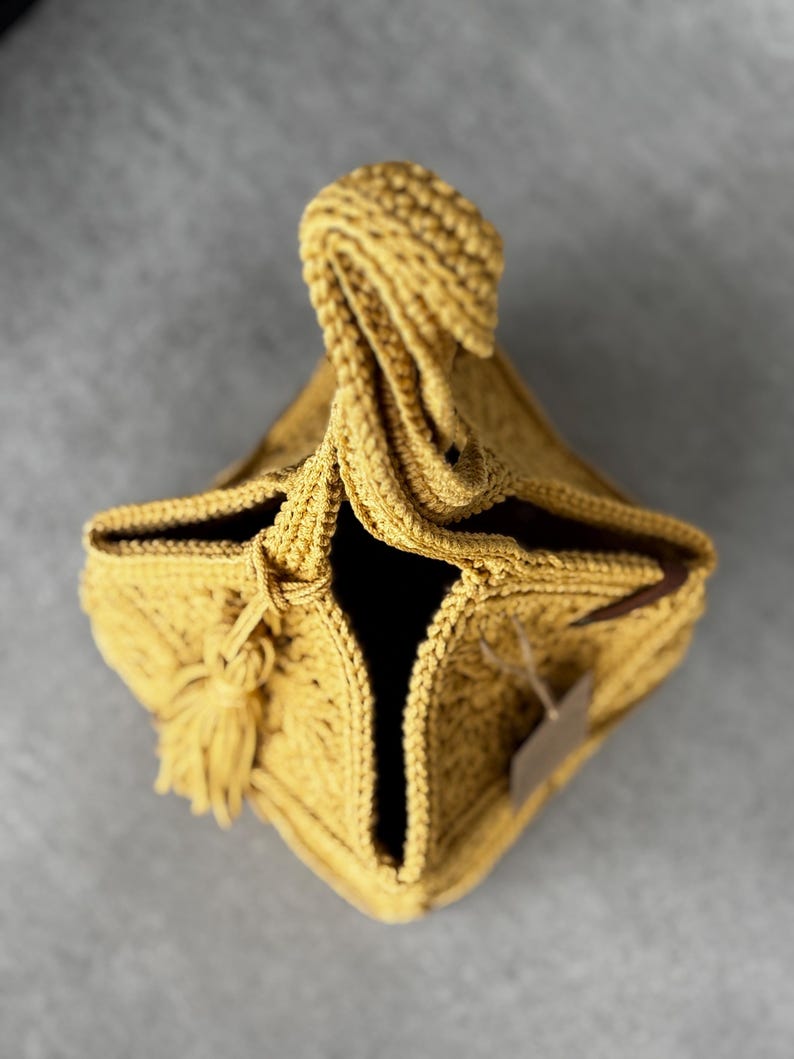 May include: Mustard yellow crocheted bag with a knotted handle and a tassel. The bag has a square base and a dark interior. A small tag is attached to the side. The bag is made of a textured yarn.