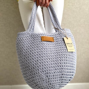 May include: A light gray crocheted tote bag with leather handles and a brown leather tag that says "Crochet Bag Lunar Cat". The bag is being held by a person wearing white pants and a yellow and blue striped shirt.