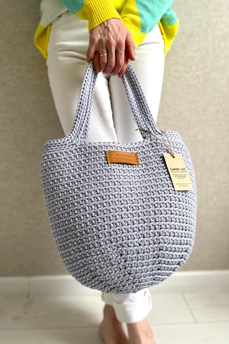 May include: A light gray crocheted tote bag with leather handles and a brown leather tag that says "Crochet Bag Lunar Cat". The bag is being held by a person wearing white pants and a yellow and blue striped shirt.