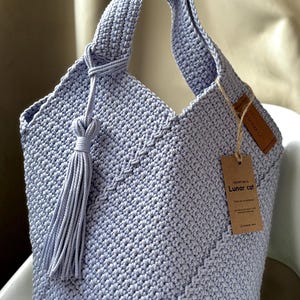 May include: A light blue crochet tote bag with a knotted handle and a long tassel. The bag features a textured stitch pattern and a small tag that reads "CROCHET BAG BY Lunar cat".