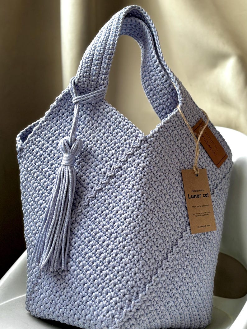 May include: A light blue crochet tote bag with a knotted handle and a long tassel. The bag features a textured stitch pattern and a small tag that reads "CROCHET BAG BY Lunar cat".