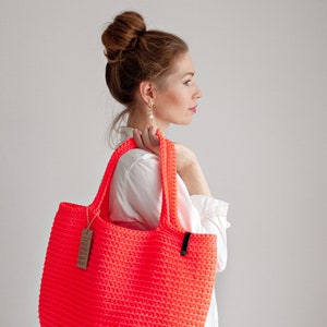 May include: A bright neon pink crocheted tote bag with black handles. The bag is being held by a person wearing a white shirt.
