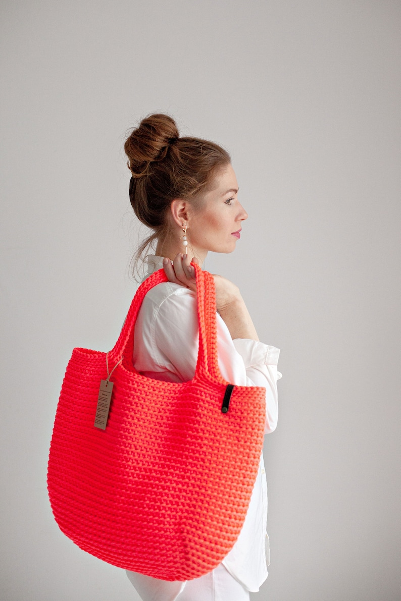 May include: A bright neon pink crocheted tote bag with black handles. The bag is being held by a person wearing a white shirt.