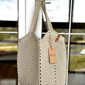 May include: A beige crochet tote bag with long handles, hanging from a white door handle. The bag has a textured, open weave design with a tag that reads "Lunar cat". The bag is made of a thick, durable yarn.
