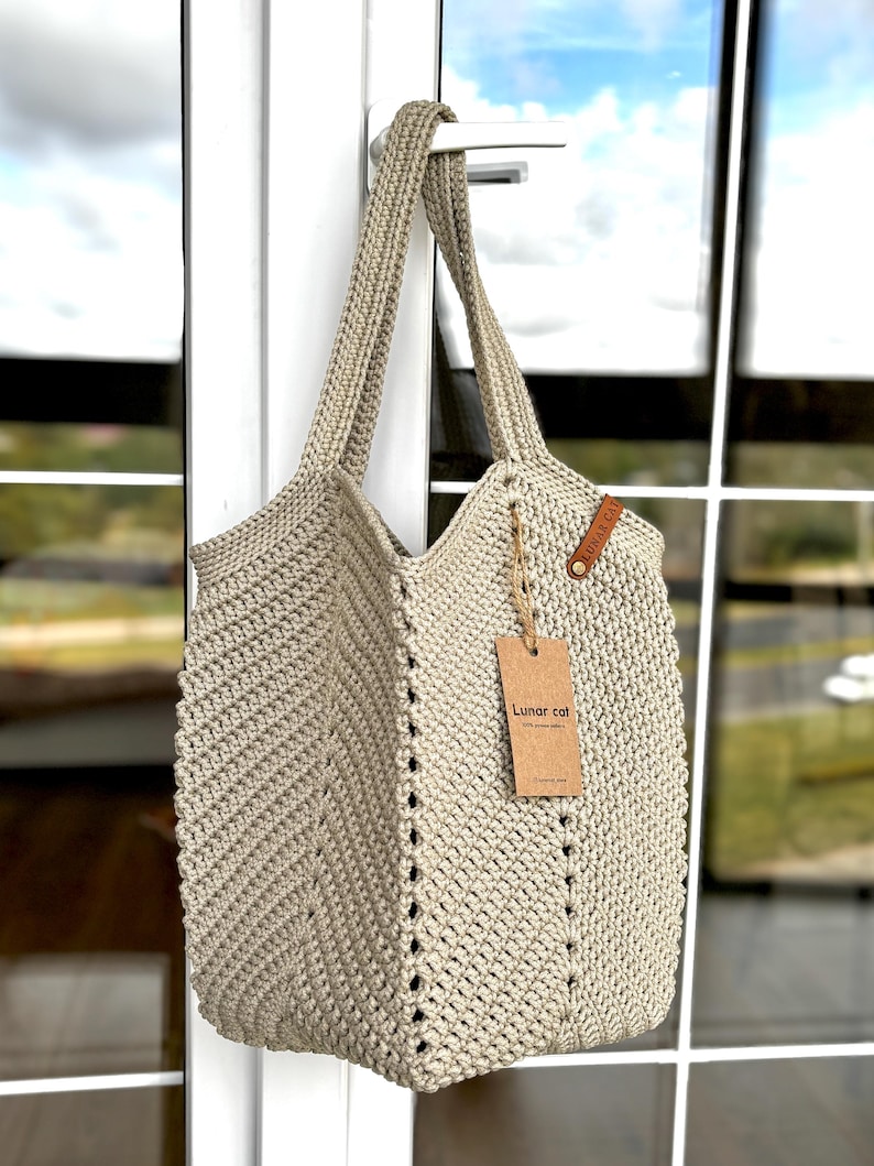 May include: A beige crochet tote bag with long handles, hanging from a white door handle. The bag has a textured, open weave design with a tag that reads "Lunar cat". The bag is made of a thick, durable yarn.