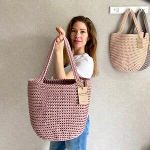 May include: A woman holds a pink crocheted tote bag with a brown tag that reads "Lunar cat". Two other crocheted tote bags in beige and gray hang on hooks in the background.