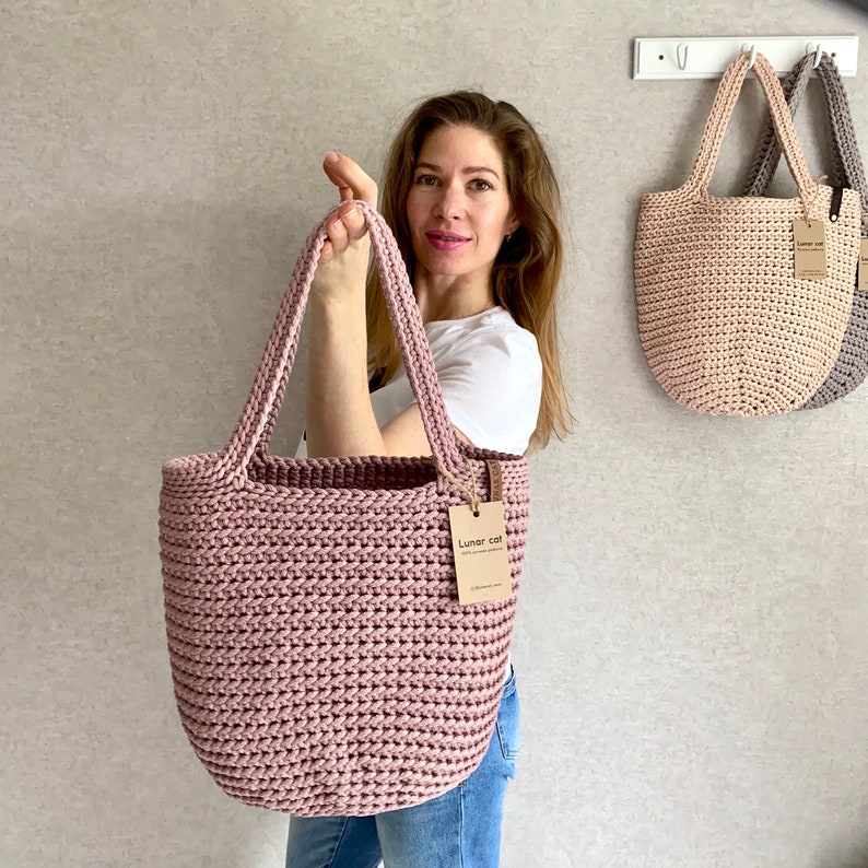 May include: A woman holds a pink crocheted tote bag with a brown tag that reads "Lunar cat". Two other crocheted tote bags in beige and gray hang on hooks in the background.