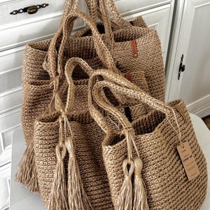 Puede incluir: Varias bolsas de mano de yute tejido de diferentes tama&ntilde;os. Las bolsas son de color beige natural con asas a juego y borlas decorativas. Una bolsa tiene una peque&ntilde;a etiqueta que dice "Lunar cat". Las bolsas se exhiben sobre una superficie blanca.