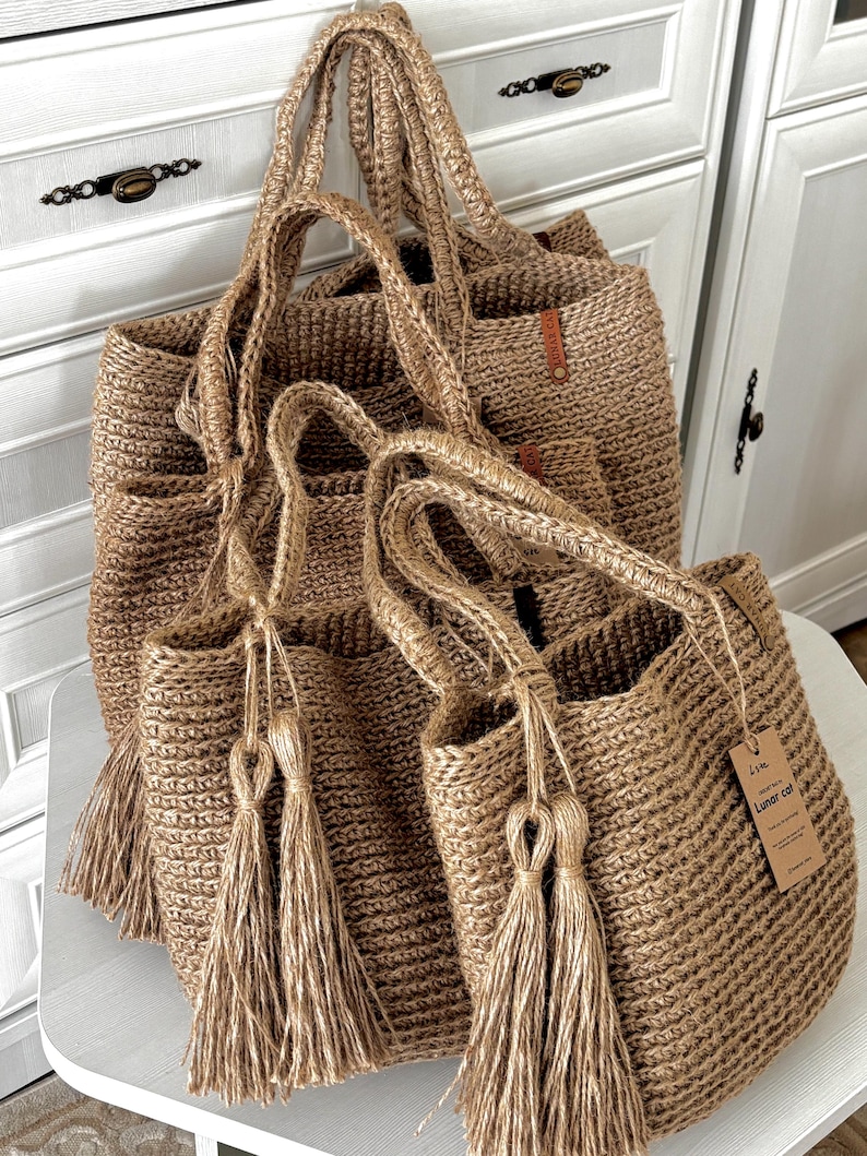 Puede incluir: Varias bolsas de mano de yute tejido de diferentes tama&ntilde;os. Las bolsas son de color beige natural con asas a juego y borlas decorativas. Una bolsa tiene una peque&ntilde;a etiqueta que dice "Lunar cat". Las bolsas se exhiben sobre una superficie blanca.