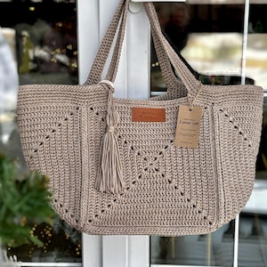 Può includere: Una borsa tote beige all'uncinetto con un motivo geometrico e un nappa. La borsa ha due manici e un'etichetta con la scritta "Lunar Cat". La borsa è appesa a una maniglia bianca.