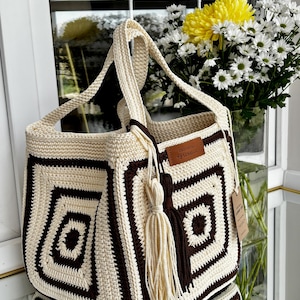 Può includere: Una borsa tote all'uncinetto di forma quadrata con un motivo geometrico di filato color crema e marrone scuro. La borsa include due manici, un'etichetta in similpelle marrone con la scritta "CROCHET by LUNAR CO" e una nappa bicolore.