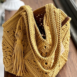 May include: Mustard yellow crochet tote bag with a geometric pattern. The bag features a long strap, a decorative tassel, and a small tag. The open top reveals the bag's interior. The bag is made of thick yarn.