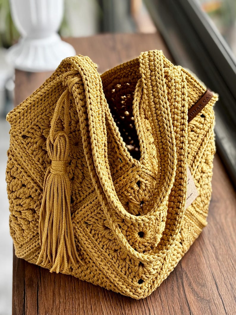 May include: Mustard yellow crochet tote bag with a geometric pattern. The bag features a long strap, a decorative tassel, and a small tag. The open top reveals the bag's interior. The bag is made of thick yarn.