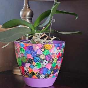 May include: A purple plant pot featuring a design of colourful dice. The pot is decorated with a variety of dice, each with a different number of sides and a different colour. The pot is sitting on a wooden surface.