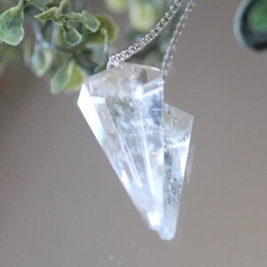 May include: A clear crystal pendant hangs from a silver chain. The pendant is shaped like a triangle with many facets. The pendant is hanging in front of a mirror with green leaves.