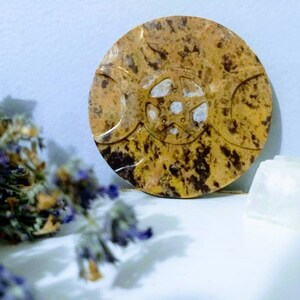May include: A round, brown and gold stone with a triple moon symbol carved into it. The stone has a speckled, marbled appearance.