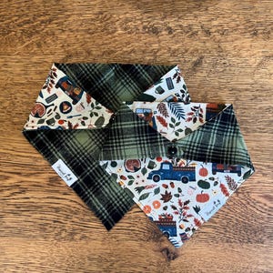 May include: A reversible pet bandana featuring a dark green and black plaid pattern on one side and a cream-colored fabric with a print of blue trucks, orange pumpkins, autumn leaves, and pie slices on the other, with a small white brand label.