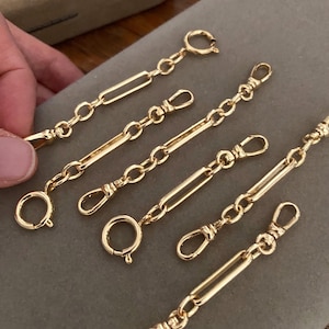 May include: Several gold-colored jewelry clasps and chains are displayed. The clasps have a spring-loaded design, and the chains feature a variety of link styles, including elongated bars and delicate chain links. The items are arranged on a neutral-toned surface.
