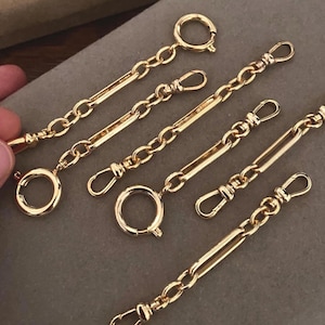 May include: Close-up of several gold-colored chain extenders with lobster claw clasps and circular clasps. The extenders are arranged on a neutral-toned surface. The chains are made of small, interconnected links.