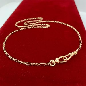 May include: A gold-toned chain necklace featuring a repeating pattern of elongated rectangular links and small circular links. The necklace includes a prominent lobster claw clasp mechanism that connects to a circular ring, creating a focal point.