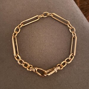 May include: A gold-colored bracelet with a mix of elongated and circular links. The bracelet features a secure clasp and is displayed on a gray surface. The jewelry piece is designed with a classic and elegant style.