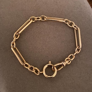 May include: A gold-colored bracelet with a chain link design. The bracelet features a series of rectangular and oval links connected by smaller circular links. A round clasp secures the bracelet. The jewelry is displayed on a gray surface.