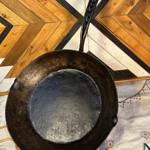 May include: A round, black cast iron skillet with a textured, dark interior surface. The pan features a long, slender handle with a braided metal design and a hook at the end, along with a small, curved helper handle on the opposite side.